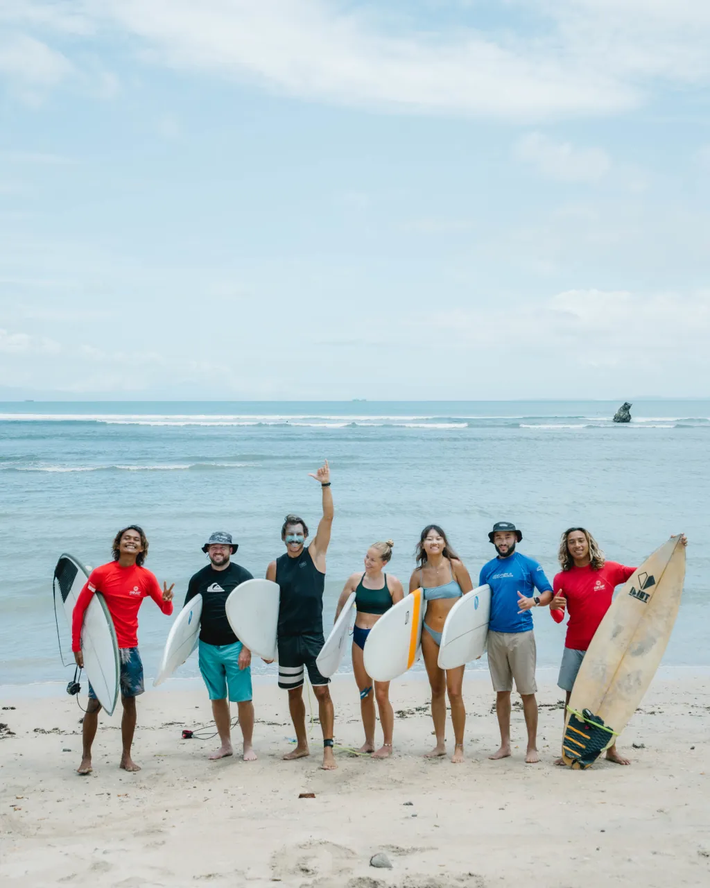a group of surfers standing side by side, holding surf boards with two surf instructors, standing on a white sand beach and smiling.