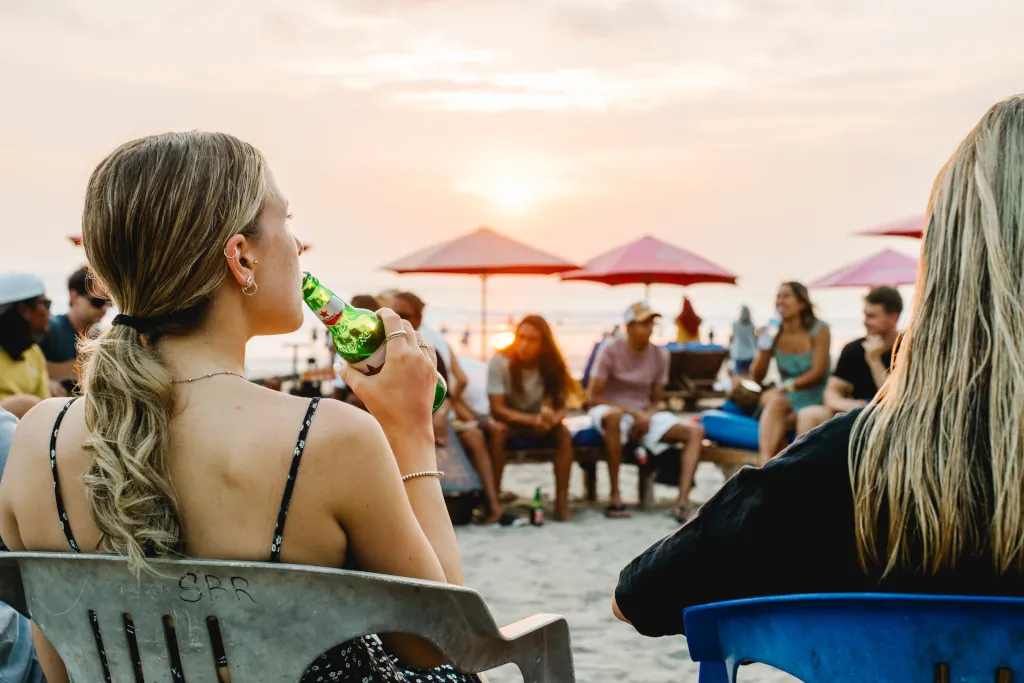 A lady drinking a bintang beer photographed from behind, sitting in a large circle of people enjoying the sunset at the beach.