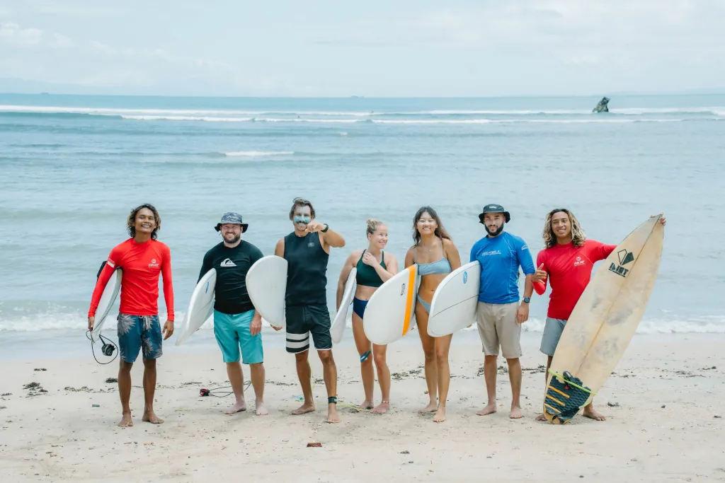 5 kima guest surfers standing holding surfboards with two surf instructors in red lycras beside them, smiling on the beach