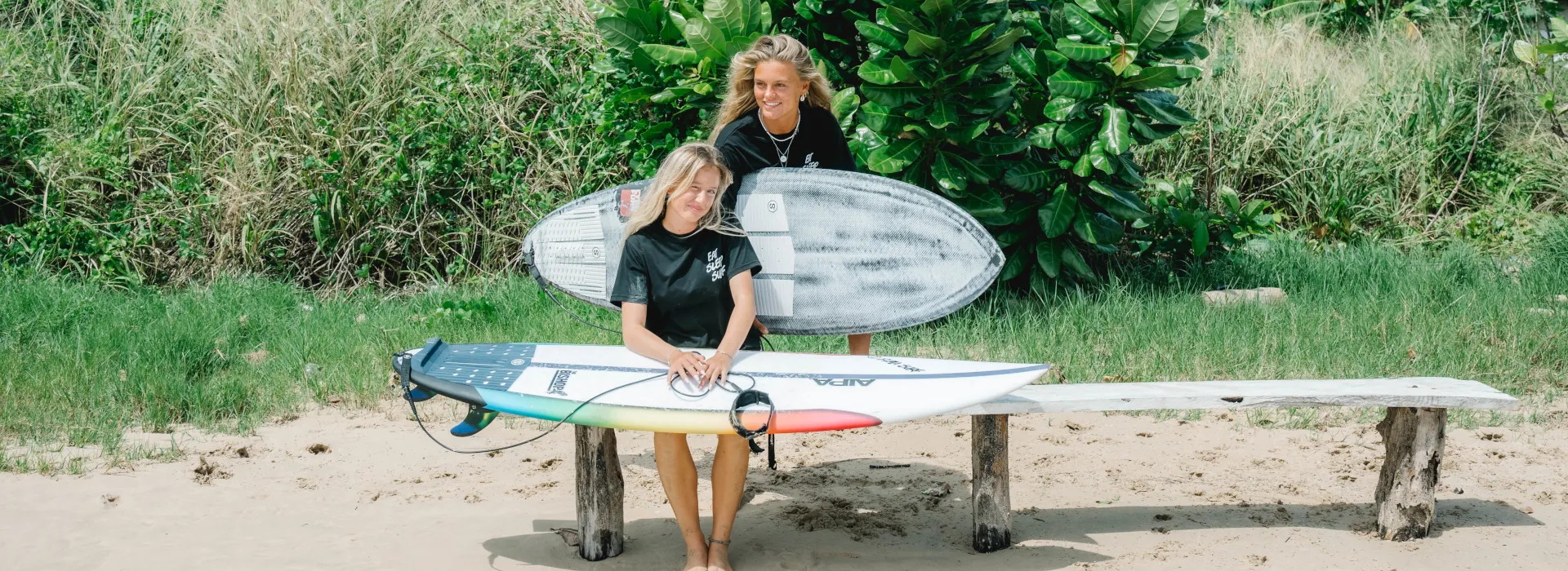2 girls sitting on a bench holding surfboards and smiling
