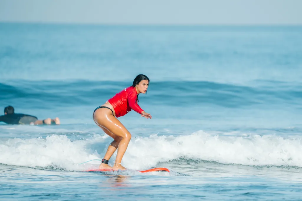 A female beginner surfer catching a white-water wave on a red foam surfboard, wearing a red lycra
