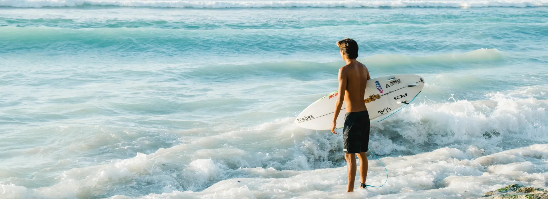 A boy holding a surfboard, looking out to the crystal-clear ocean on a bright and sunny day.