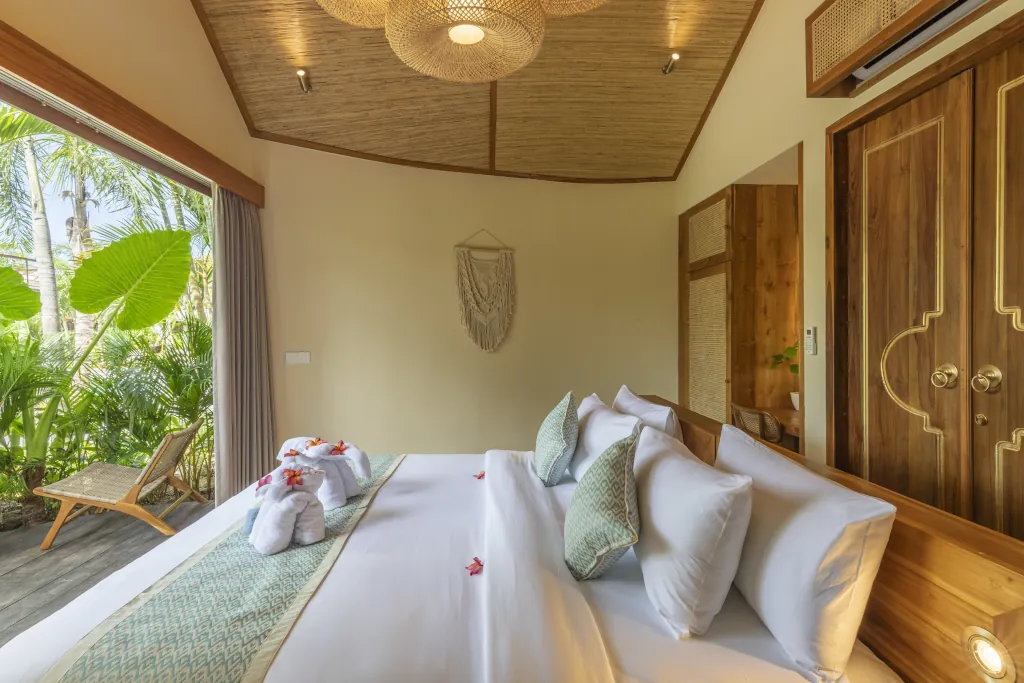 A room taken on a side angle in Uluwatu, with white bed sheets, a blue throw over and decorative pillows and flowers as well as towel art on the bed