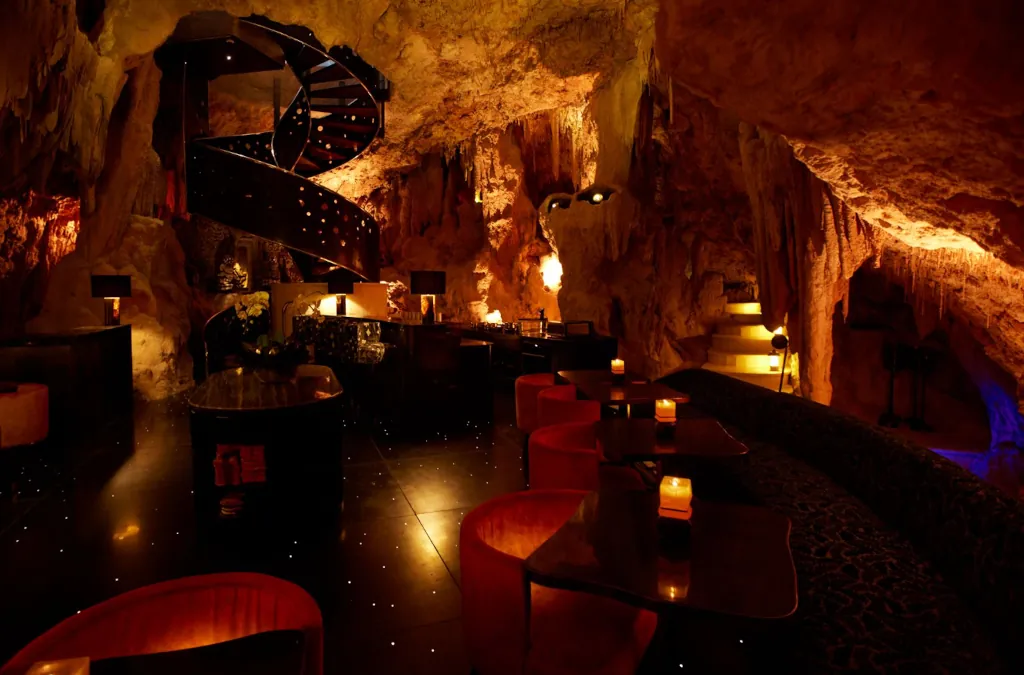 a candle lit natural limestone cave with dining tables and a spiral staircase.