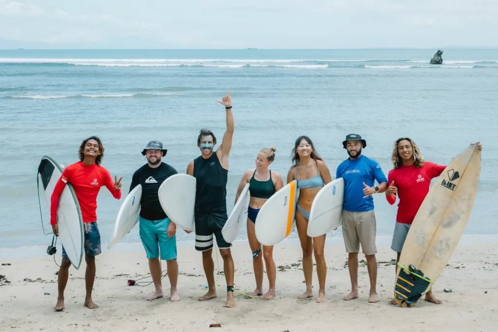 Surfers standing on the beach holding their surfboards, with 2 surf guides by them, all smiling at the camera