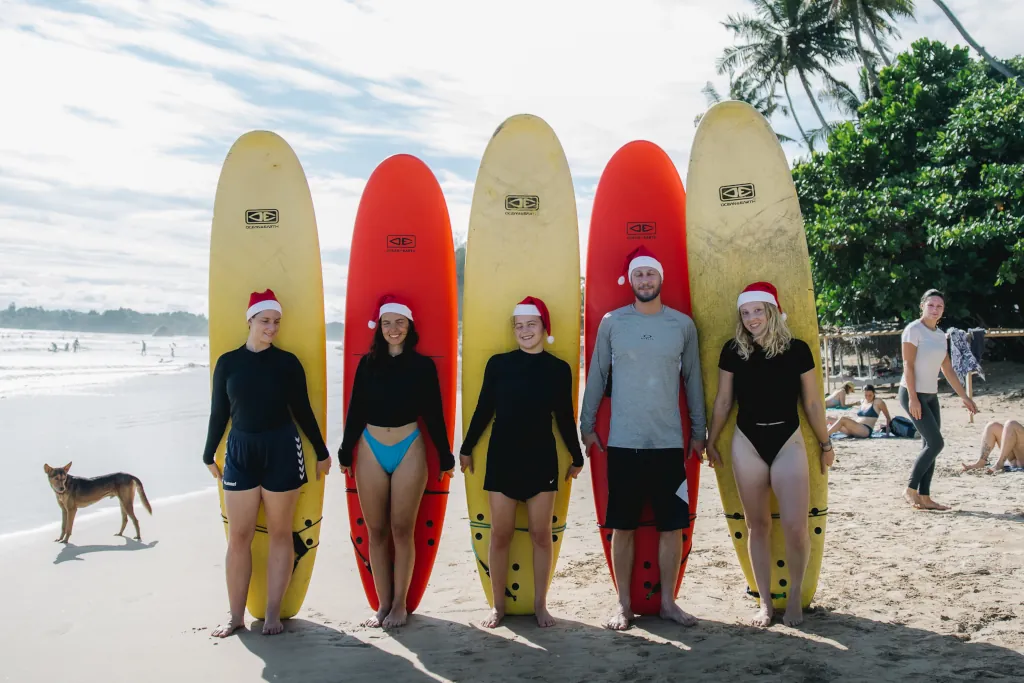 5 GUESTS STANDING ON THE BEACH WITH CHRISTMAS HATS AND SOFT TOP SURFBOARDS