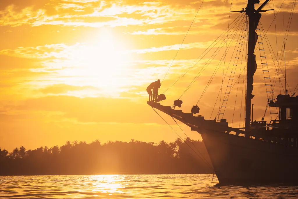 A sunset in the background, with two people on the bow of a sailing boat
