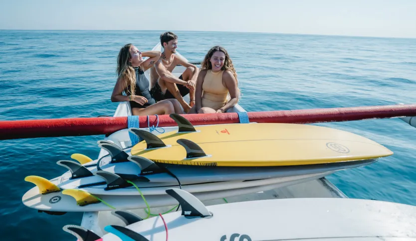 Two girls and one boy smiling at the front of the boat with surfboards stacked in front of them and the blue ocean in the background.