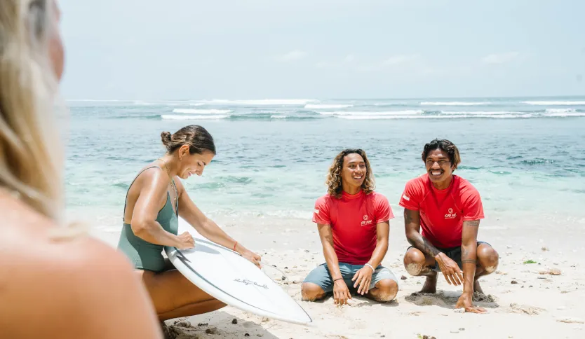 A girl waxing her surfboard with two surf instructors kneeling in the sand and smiling, with the clear ocean in the background.