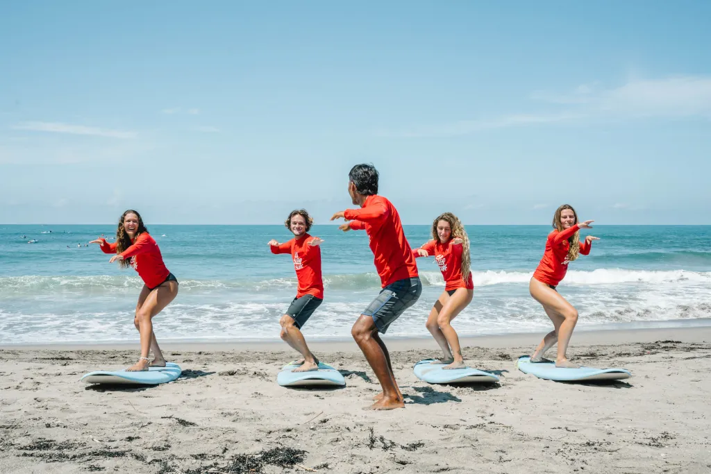 4 students learning to surf on the beach front in Bali, the surf guide is in front with his back to the camera and showing the surf stance.