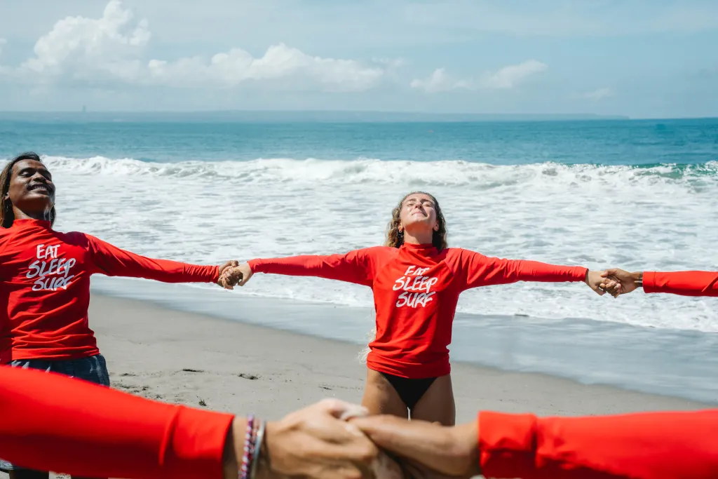 Guests learning to surf and their surf guides, holding hands and stretching on the beach.