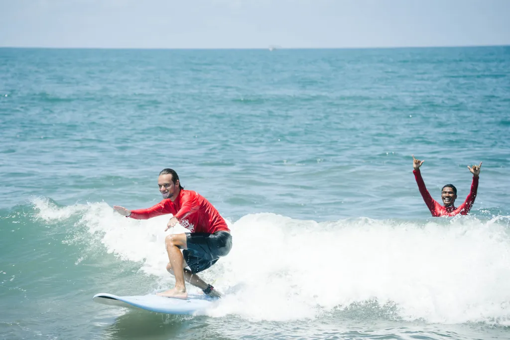 A male surfer catching a wave on a blue foambaord with the surf guide cheering him on from behind.