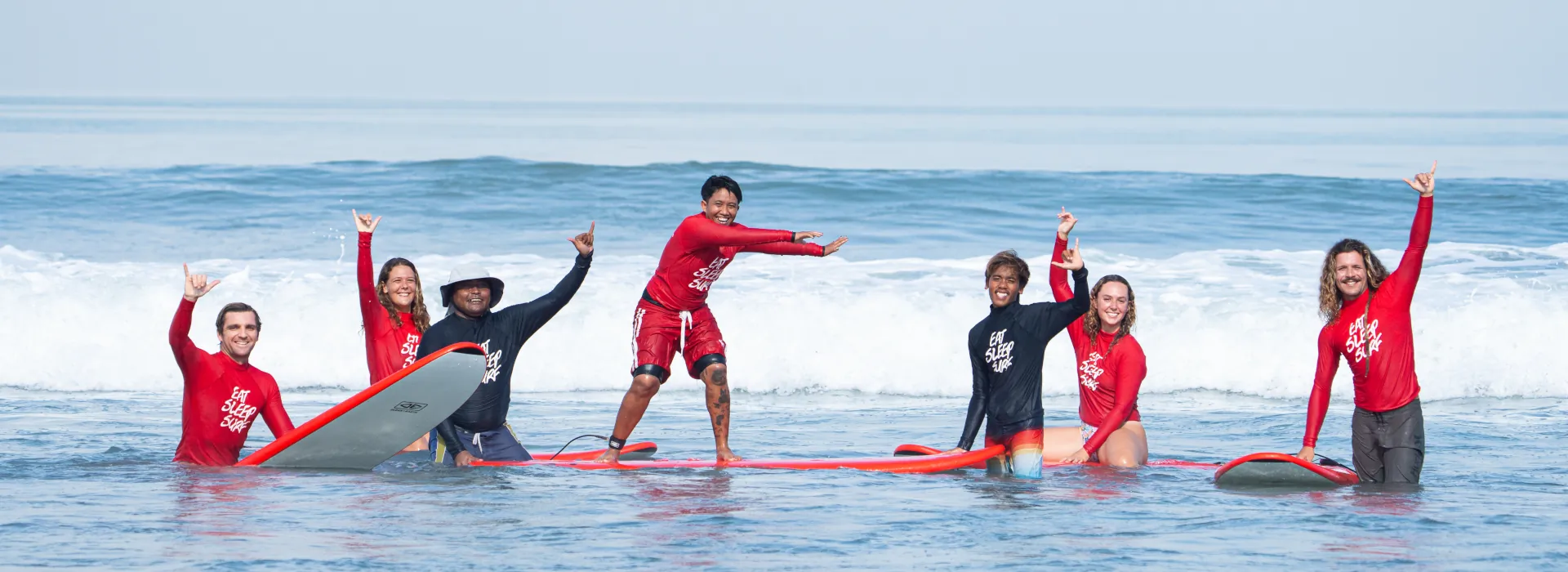 5 GUESTS LEARNING TO SURF WITH 2 SURF GUIDES FROM KIMA SURF SCHOOL, THE GROUP IS POSING IN THE OCEAN ON THEIR SURFBOARDS.